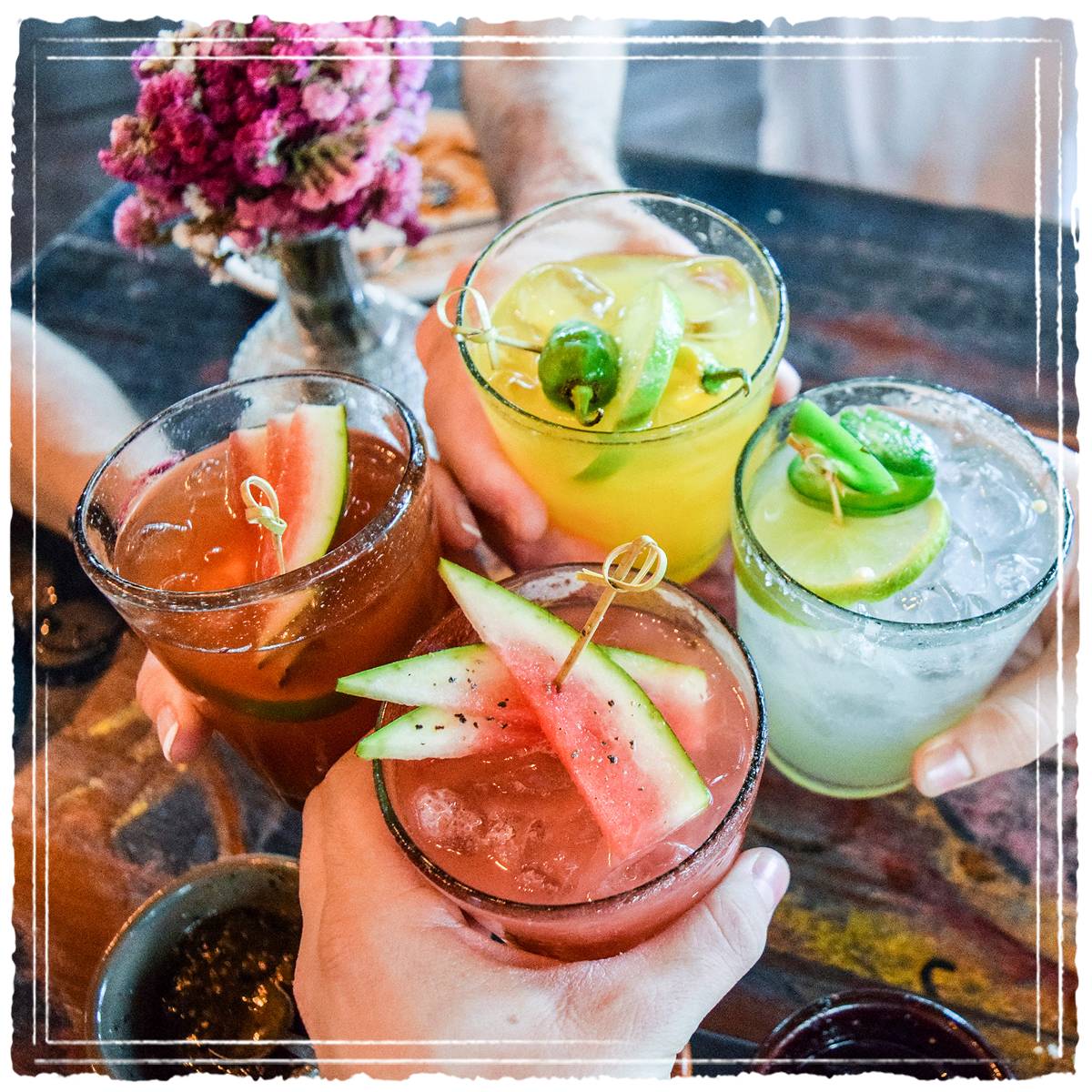 Four cocktails being cheers-ed featuring colorful garnishes like watermelon, lime and jalapeno.