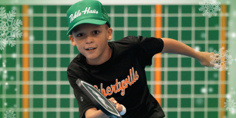 Young boy wearing green Pickle Haus hat playing pickleball with winter snowflake border around image.